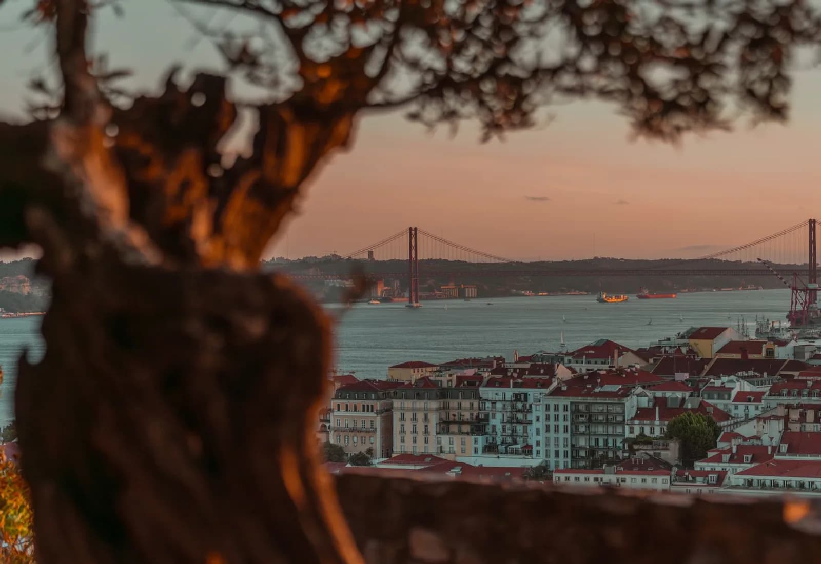 Beautiful sunset over Lisbon with a view of the iconic 25 de Abril Bridge, framed by trees.